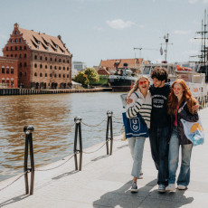 A group of three smiling young adults walk arm in arm along a sunny waterfront in Gdańsk, Poland. Behind them, historic brick buildings, including the Polish Baltic Philharmonic, line the Motława River, with moored ships and a clear blue sky completing the picturesque scene. The students, casually dressed and carrying University of Gdańsk tote bags, exude a sense of camaraderie and joy as they explore the city.